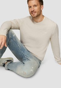 Lichtbeige gebreide trui met geribbelde boorden, gecombineerd met vervaagde blauwe jeans met versleten accenten en een verfspattenontwerp.