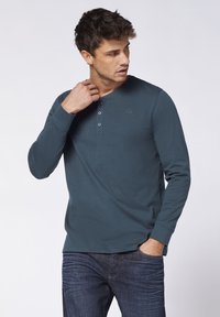 Langarm-Henley-Shirt in dunklem Teal, aus weichem Stoff, mit einer Knopfleiste mit drei Knöpfen und einem dezenten gestickten Logo auf der Brust.