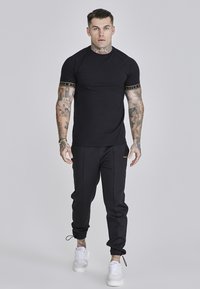 Sort tætsiddende t-shirt med guld logo detaljer på ærmerne, kombineret med sorte joggers og hvide sneakers, hvilket skaber et sporty look.