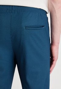 Turkusowe prążkowane spodnie mają elastyczny pas, tylną kieszeń naszywaną oraz gładką fakturę. Projekt podkreśla dopasowany krój.