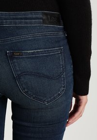 Jeans de denim azul oscuro con una textura suave, que presentan un parche de cuero de la marca en la cinturilla y un diseño de bolsillo trasero curvado.