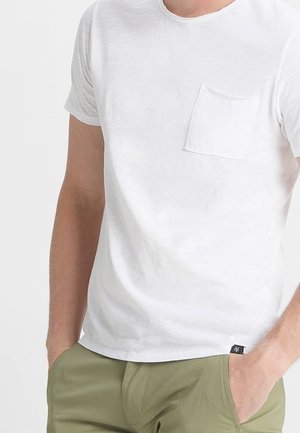 Man draagt een effen wit T-shirt met korte mouwen en een borstzak, en olijfgroene broek, handen in de zakken.