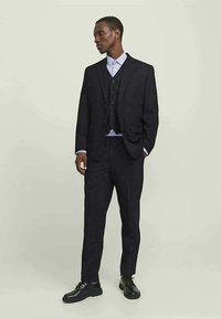 Costume sombre comprenant une veste sur mesure et un gilet, avec un pantalon assorti. L'ensemble est complété par une chemise bleu clair et des chaussures noires.
