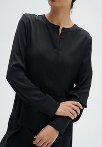 Svart långärmad blus med rund halsringning, knappstängning och slät textur. Designen har skräddarsydda ärmar och en avslappnad passform.