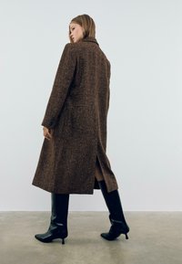 Manteau en tissu pied-de-coq brun avec un col cranté, des manches longues et une fente sur le côté, associé à des bottes noires jusqu'aux genoux sur un fond neutre.