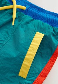 Shorts de bain bleus, verts sarcelle et rouges avec un détail de poche verticale jaune et un cordon jaune sur une ceinture élastique bleue.
