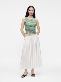Jeune femme aux longs cheveux foncés portant un haut vert à motifs sans manches, une jupe midi blanche et des sandales noires ouvertes à l’avant, debout les mains derrière le dos.