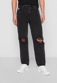 Black denim jeans with ripped knee details, relaxed fit, five-pocket design, and a logo patch on the front. Worn with white athletic shoes.