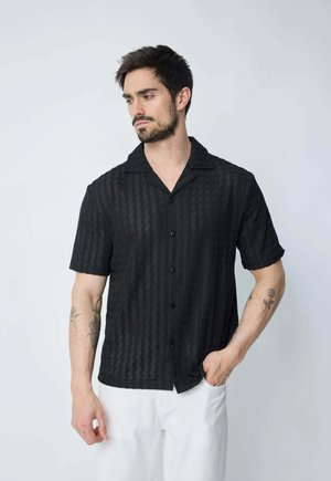 Chemise noire à manches courtes avec un motif texturé, col et boutons. Portée avec un pantalon blanc. Teint de peau visible sur la main avec un tatouage.