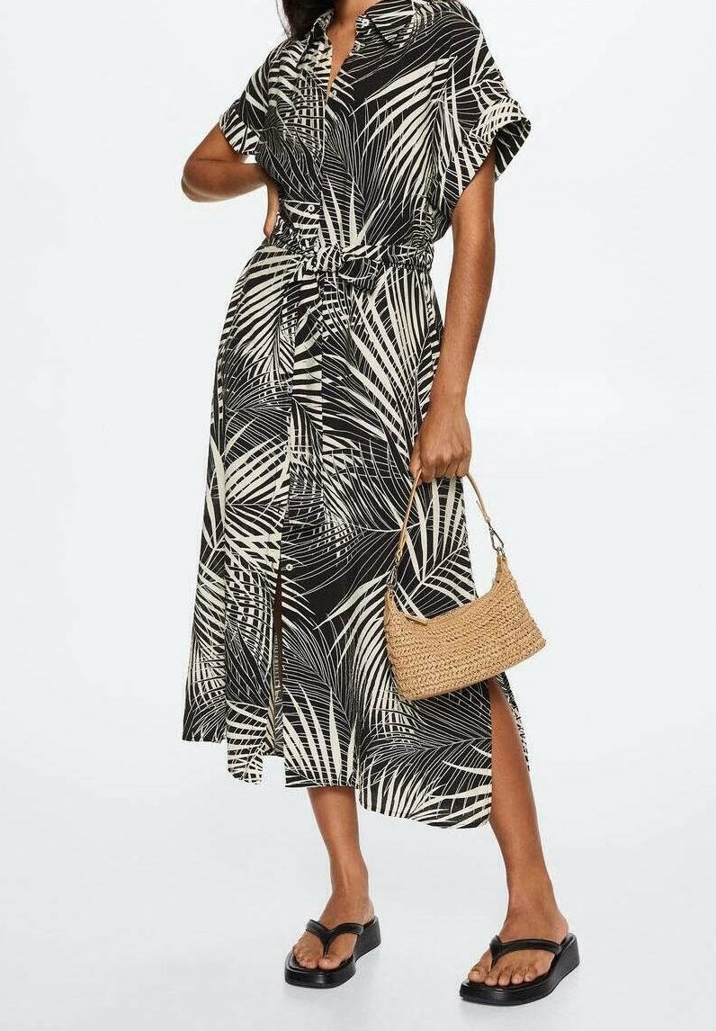 Femme portant une robe chemise midi imprimée feuilles de palmier noir et blanc, tenant un sac à main en paille tressée, avec des sandales compensées noires.