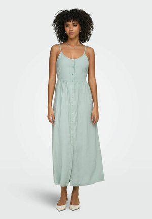 Femme aux cheveux bouclés portant une robe maxi sans manches vert clair à boutons sur le devant et des talons blancs, debout devant un fond uni.