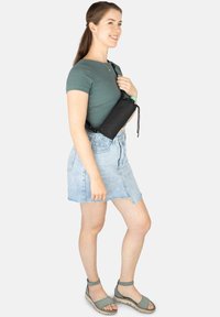 Schwarze Umhängetasche mit Kordelzugverschluss, getragen zu einem grünen Oberteil und einem hellblauen Denim-Rock, kombiniert mit hellblauen Sandalen.