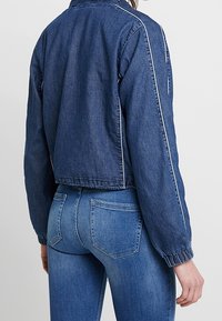 Blå denimjacka med en kortad design, elastiska ärmar och synlig sömnad. Bärs över blå jeans, vilket ger en avslappnad passform.