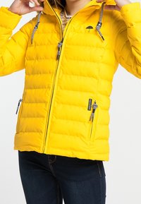 Gul vadderad jacka med dragkedja framtill, svarta metallaccenter, elastiska dragsnören och två fickor med dragkedja på sidan. Mjuk textur, varm design.