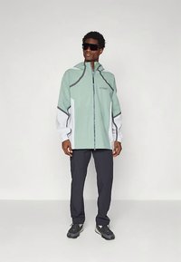 Casaco impermeável verde claro com detalhes em cinza e fecho de correr, combinado com calças escuras e sapatos pretos, usado por uma pessoa com óculos de sol.