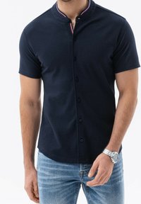 Polo de manga corta en color azul marino, con cuello alzado, cierre de botones y ribete del cuello en contraste. Textura suave, corte entallado, con detalles de costura visibles.