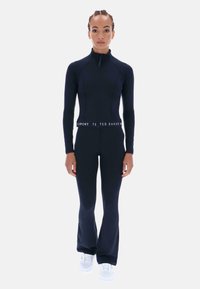 Čierny atletický body s dlhými rukávmi, vysokým golierom a zipsovým zapínaním. Rozšírený dizajn nohavíc s logovaným pásom. Viditeľné biele tenisky.