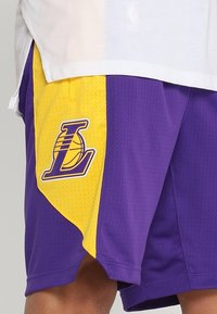 Violetit ja keltaiset koripalloshortsit, jotka on valmistettu verkkomateriaalista ja joissa on brodeerattu logo vasemmalla jalalla. Suunnittelussa on kontrastisia värejä ja tekstuureja.