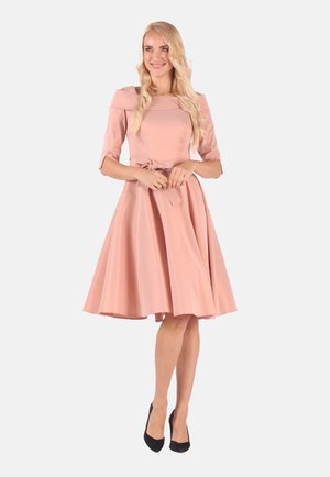 Blonde woman in knee-length pink dress with belt and three-quarter sleeves, wearing black heels, standing against a plain light background.