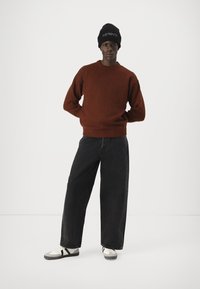 Pull en maille marron, jeans noirs à coupe large, et baskets blanches avec des accents noirs. Bonnet noir avec du texte. Tenue simple et décontractée.