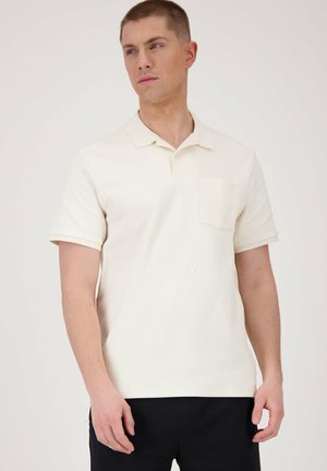 Valkoinen lyhythihainen polo-paita, jossa on kaulus, teksturoitu kangas ja rintatasku, käytetty tummien housujen kanssa. Yksinkertainen muotoilu ja neutraali väri.