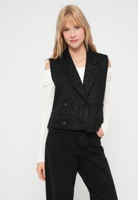 Crni, teksturirani dvoredni prsluk s otvođenim ovratnikom, sadrži dva prednja džepa i velike dugmadi, nosi se preko svijetlo bež majice.