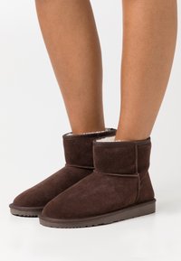 Brown suede ankle boots with a soft white lining, flat soles, and a rounded toe design. Features a stitched seam and a cuffed opening.