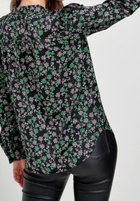 Schwarze Bluse mit floralen Mustern in Weiß und Grün, langen Ärmeln und einem leicht abgerundeten Saum. Hergestellt aus leichtem Material.