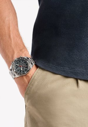 Silver metal watch with a black dial, three sub-dials, and white accents. Worn on a hand, with dark polo shirt and beige pants.