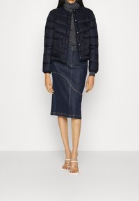 Žena v černé prošívané bundě, šedém roláku, tmavě modré midi sukni z denimu a bílých otevřených lodičkách stojí proti bílé zdi.