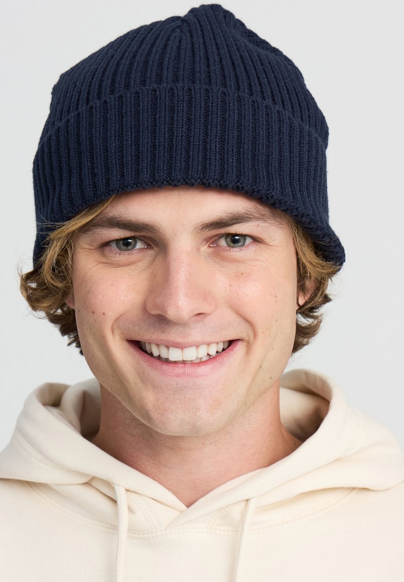 Beanie a coste navy con una texture morbida, caratterizzato da una vestibilità aderente e un bordo ripiegato, indossato sopra una felpa chiara.