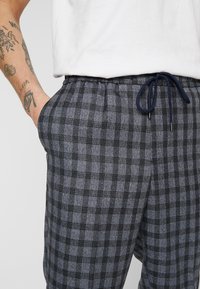 Männer Plaid Lounge-Hosen in Grau und Schwarz. Elastischer Bund mit marineblauem Kordelzug, Seitentaschen und weichem Textil. 