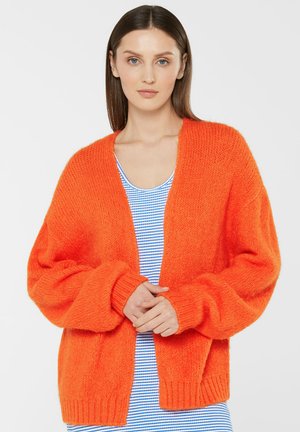 Frau trägt einen locker sitzenden, leuchtend orangefarbenen, gestrickten Cardigan über einem blau-weiß gestreiften Top, die Hände vor dem Körper verschränkt, neutraler grauer Hintergrund.