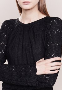 Top in maglia nera con maniche lunghe testurizzate che presentano un motivo intricato. Il collo è rotondo e il tessuto ha una lieve lucentezza.