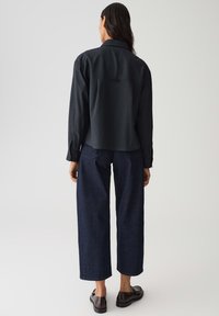 Donna in piedi di spalle con indosso una maglietta nera a maniche lunghe, jeans larghi blu scuro e mocassini in pelle nera su uno sfondo bianco.