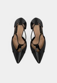 Μαύρα γυναικεία φλατ παπούτσια με μυτερή μύτη, ταινία Τ και πόρπη στον αστράγαλο, σε μπεζ εσωτερική σόλα, εμφανιζόμενα από πάνω σε λευκό φόντο.