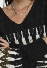 Schwarz-weißer gestrickter Pullover mit V-Ausschnitt, der ein strukturiertes Muster und weite Ärmel aufweist. Eine goldene Schlangenkette ist sichtbar.