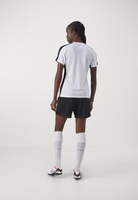Hvit idrettsskjorte med svarte sideaksenter, svarte shortser, hvite kne-høye sokker og fotballsko med røde detaljer, sett fra baksiden.