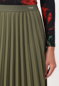 Gonna plissettata verde oliva con una finitura testurizzata e una vita ampia. Presenta il marchio 'HILFIGER NEW YORK' in vita.