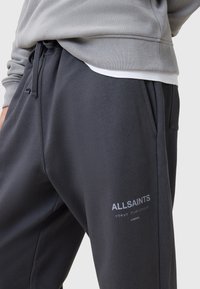 Zwarte joggingsbroek van een gladde stof, met een elastische tailleband en zijzakken. "ALLSAINTS" logo in het wit gedrukt op het onderbeen.