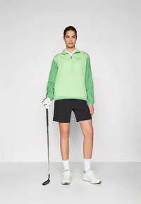 Jachetă verde deschis cu fermoar până la piept și mâneci lungi, pantaloni scurți negri, șosete albe și încălțăminte de golf, ținând un crosă de golf. Material neted, ușor.