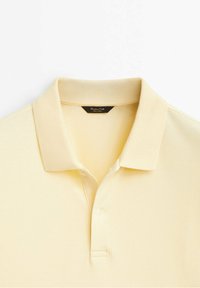 Polo jaune clair à manches courtes avec un col plié et deux boutons sous le col, posé à plat sur un fond blanc.