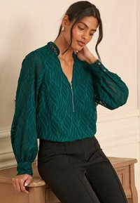 Blusa teal con motivo di foglie testurizzate, collo con zip e lunghe maniche trasparenti, abbinata a pantaloni neri aderenti, su uno sfondo neutro.