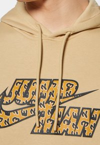 Sweat à capuche beige avec cordons, grand logo Nike et texte "JUST WIN" en jaune avec motif léopard noir.