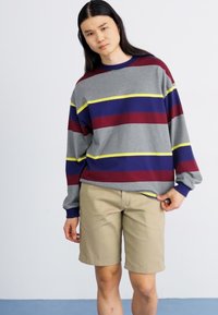 Chemise grise à manches longues avec des rayures horizontales bordeaux, bleues et jaunes, associée à un short beige, se tenant devant un fond uni.