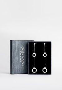 Orecchini a cerchio argentati con goccia, presentati in una elegante scatola regalo foderata in velluto nero, caratterizzati da anelli circolari su catene e una finitura argentata lucida.