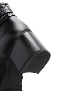 Chaussure en cuir noir avec une bride de cheville texturée, un talon carré avec une finition lisse et un design de bout rond.
