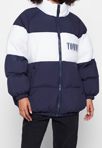 Person som bär en marinblå och vit fuskpälsjacka med dragkedja framtill och "Tommy Jeans"-logotyp på bröstet, tillsammans med svarta byxor.
