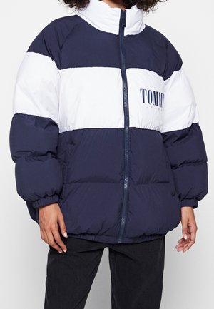 Osoba nosząca granatową i białą kurtkę puchową z zamkiem błyskawicznym z przodu oraz logo "Tommy Jeans" na klatce piersiowej, w zestawie z czarnymi spodniami.