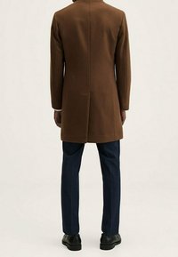 Cappotto in lana marrone con taglio dritto, con uno spacco sul retro, indossato sopra pantaloni a quadri blu navy e scarpe nere. Texture liscia.